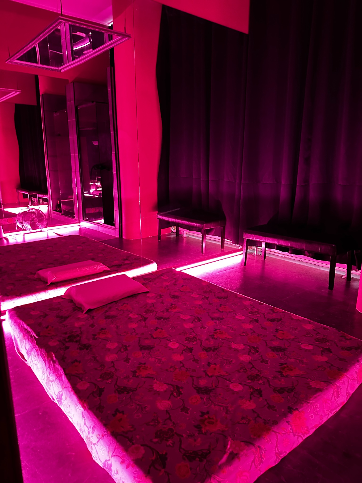 Dwa łóżka z kwiatowym wzorem i poduszkami w pomieszczeniu oświetlonym różowym neonem, z czarnymi zasłonami i ławkami w tle.