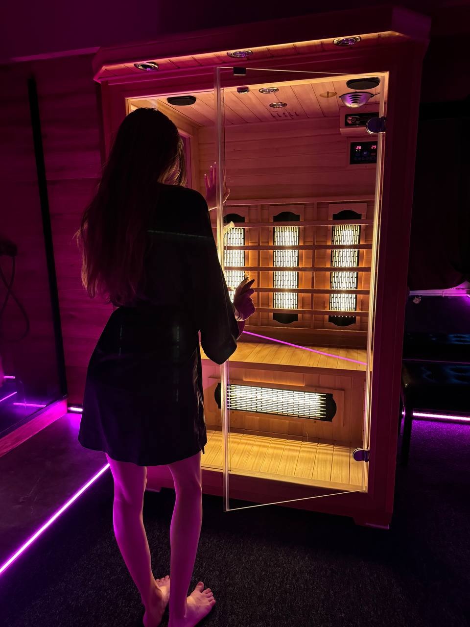 Kobieta w szlafroku stoi przed otwartą sauną infrared z drewnianym wnętrzem, oświetloną ciepłym światłem, w pomieszczeniu z fioletowym oświetleniem LED.