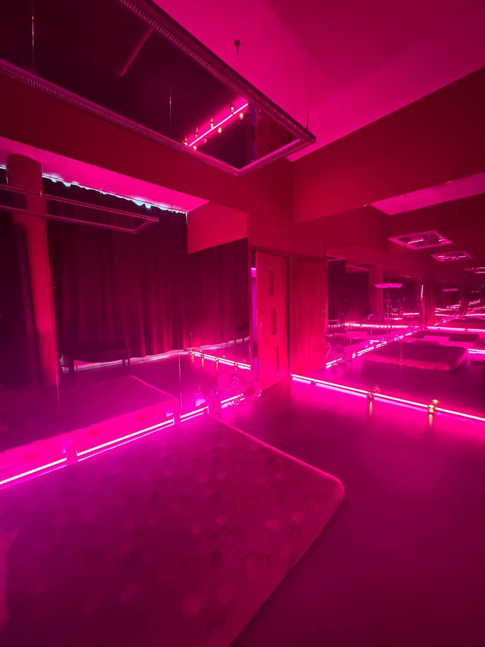 Wnętrze salonu z łóżkami do masażu, oświetlone intensywnym różowym światłem neonowym, z lustrzanymi ścianami i sufitem.