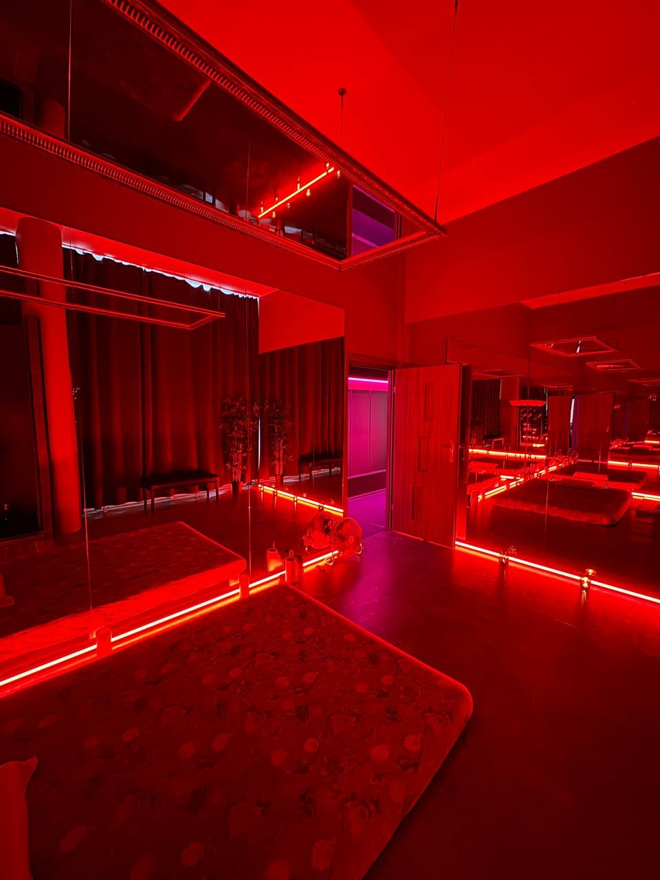 Sala do masażu z czerwonym oświetleniem LED, lustrami na ścianach i suficie, oraz dwoma wentylatorami stojącymi obok siebie.
