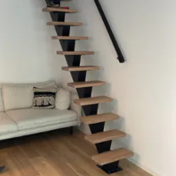 Minimalistyczne schody z drewnianymi stopniami i czarną, metalową konstrukcją, umieszczone przy białej ścianie w nowoczesnym wnętrzu z jasną podłogą i kremową sofą.