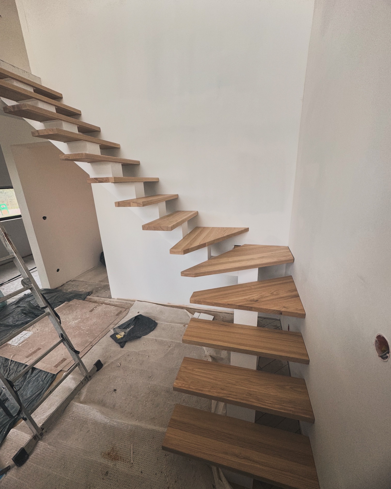 Drewniane schody wspornikowe o trójkątnych stopniach, zamontowane przy białej ścianie podczas remontu, widoczne zabezpieczenia podłogi i drabina.
