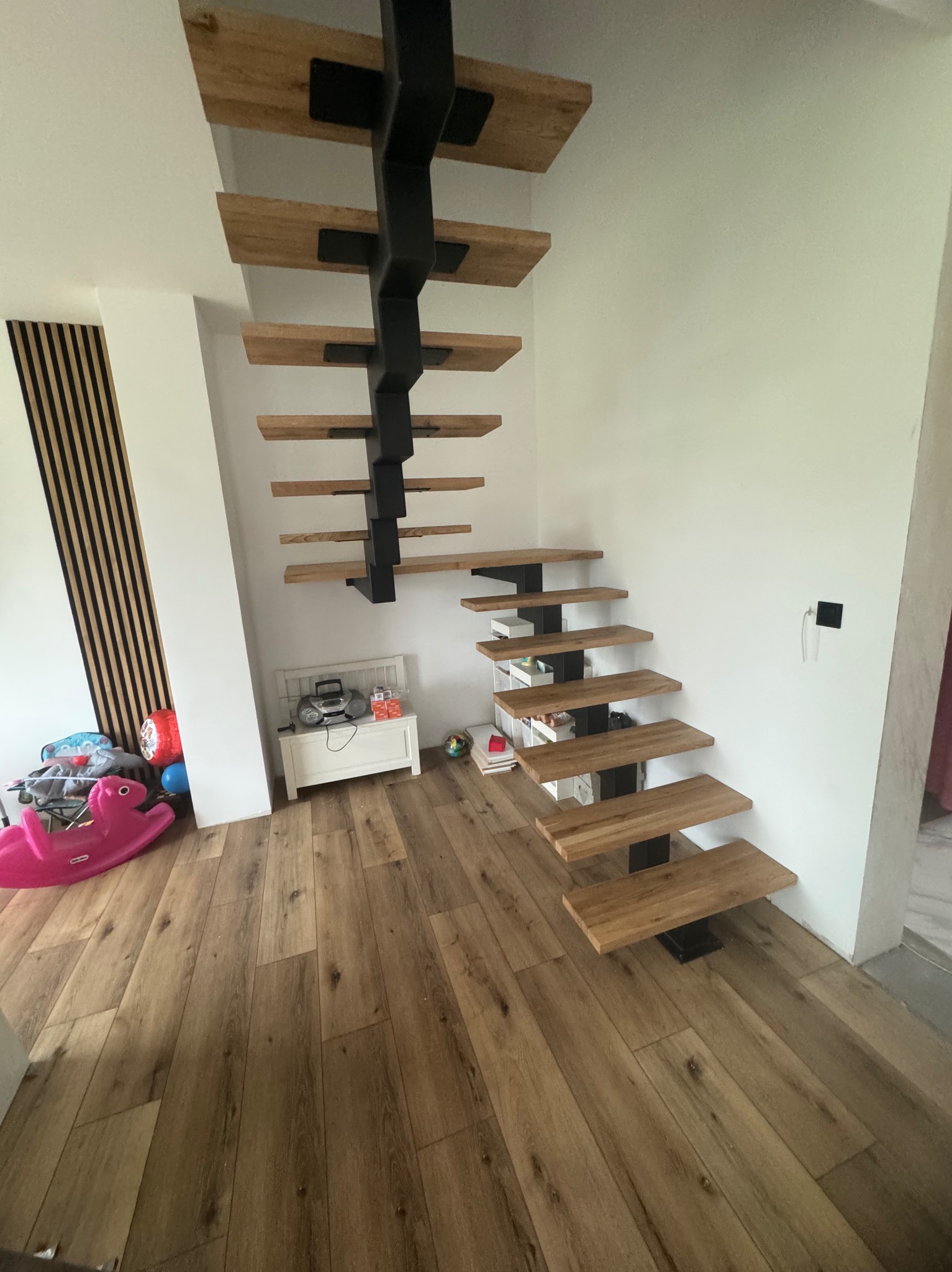 Nowoczesne schody wspornikowe z drewnianymi stopniami i czarną metalową konstrukcją nośną, widok z dołu na tle jasnego wnętrza z drewnianą podłogą i zabawkami w rogu