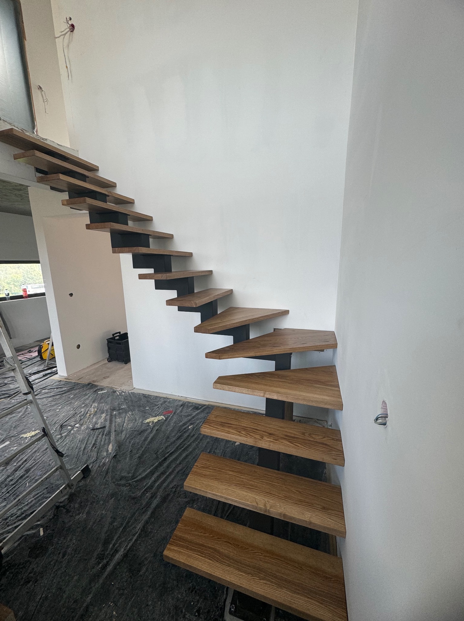 Drewniane stopnie schodów na metalowej konstrukcji, zamontowane przy białej ścianie w surowym wnętrzu, zabezpieczone folią malarską na podłodze, widoczna drabina.