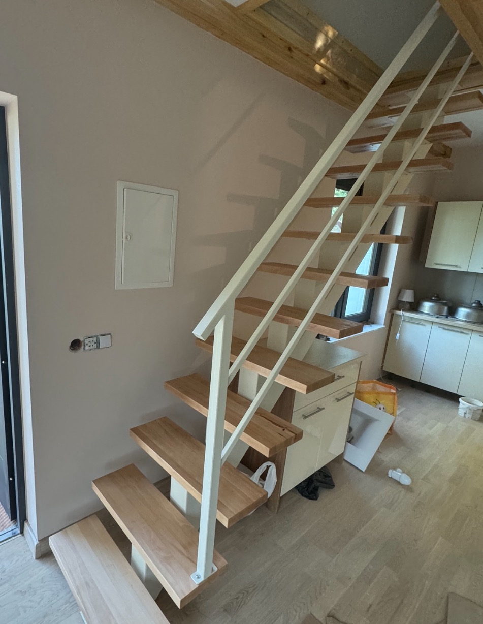 Nowoczesne schody metalowe z drewnianymi stopniami i białą balustradą w jasnym wnętrzu domu, widoczne elementy wykończeniowe.