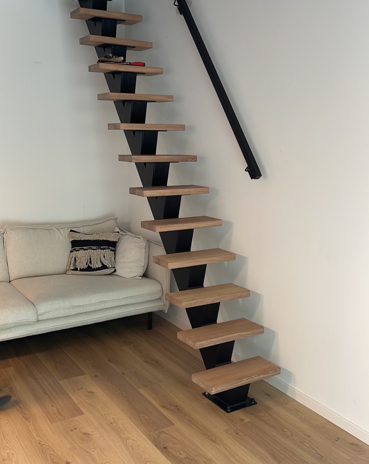 Minimalistyczne schody z drewnianymi stopniami i czarną, metalową konstrukcją, umieszczone przy białej ścianie w nowoczesnym wnętrzu z jasną podłogą i kremową sofą.