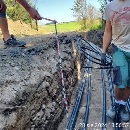WM WOJCIECH KULIK - Wąski wykop w ziemi z ułożonymi rurami, obok pracownik z łatą mierniczą, w tle zielone pola.