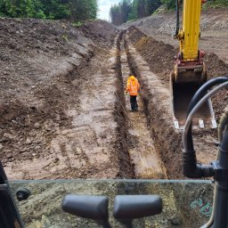 WM WOJCIECH KULIK - Wykopane równoległe rowy w gruncie, widok z kabiny koparki, pracownik w odblaskowej kamizelce kontroluje głębokość wykopu.