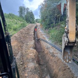 WM WOJCIECH KULIK - Głęboki wykop w ziemi pod rurę, widok z kabiny koparki, pracownicy w pomarańczowych kombinezonach w oddali, teren zalesiony