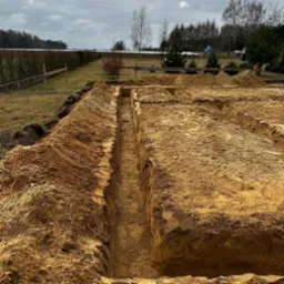 Wykopane wąskie rowy pod fundamenty na działce z widocznym ogrodzeniem i drzewami w tle, ziemia piaszczysta, pochmurne niebo.