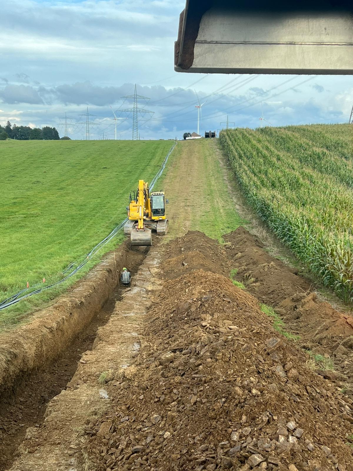 Żółta koparka w trakcie wykonywania wykopu pod instalacje na polu uprawnym, widoczny pracownik w kasku i flanelowej koszuli w wykopie oraz rury wzdłuż wykopu, w tle słupy wysokiego napięcia...