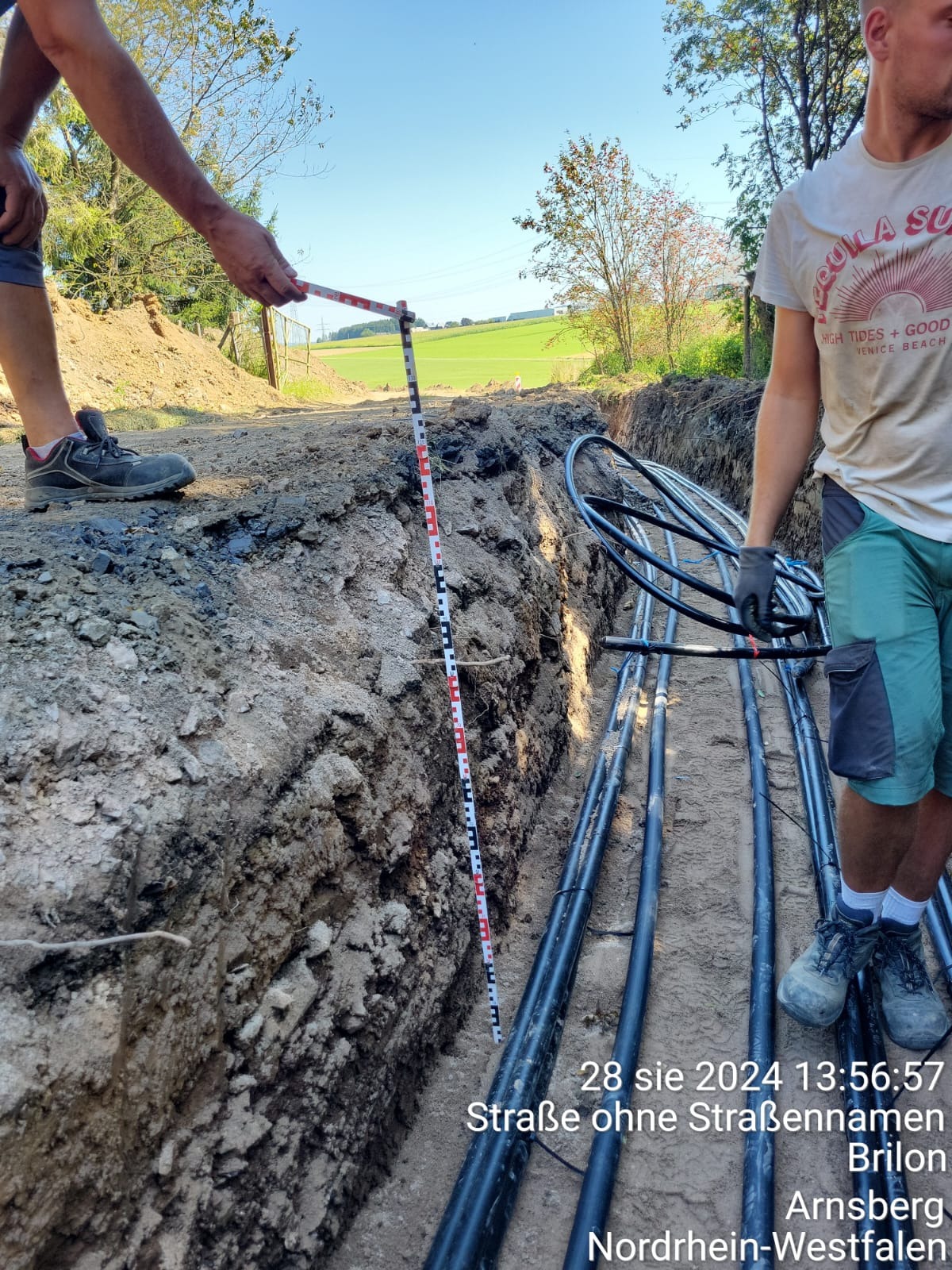 Wąski wykop w ziemi z ułożonymi rurami, obok pracownik z łatą mierniczą, w tle zielone pola.