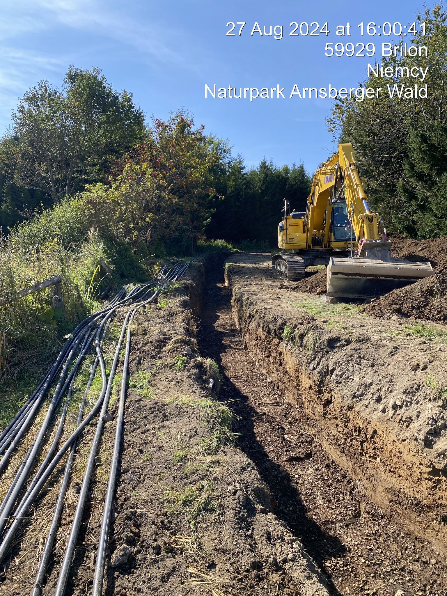 Żółta koparka Komatsu podczas wykopu wąskiego rowu w ziemi, obok leżą rury instalacyjne, widok na teren Naturpark Arnsberger Wald, Niemcy.