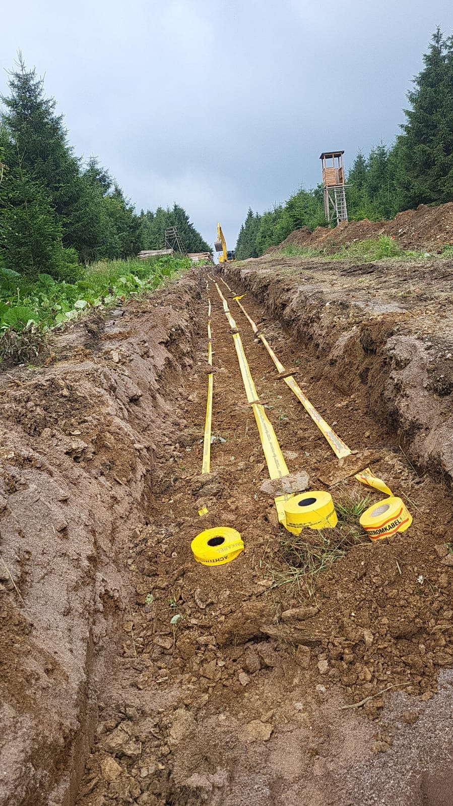 Wąski wykop w ziemi z ułożonymi rolkami taśmy ostrzegawczej 'ACHTUNG STROMKABEL' wzdłuż trasy, w tle las i wieża obserwacyjna, widoczna koparka.