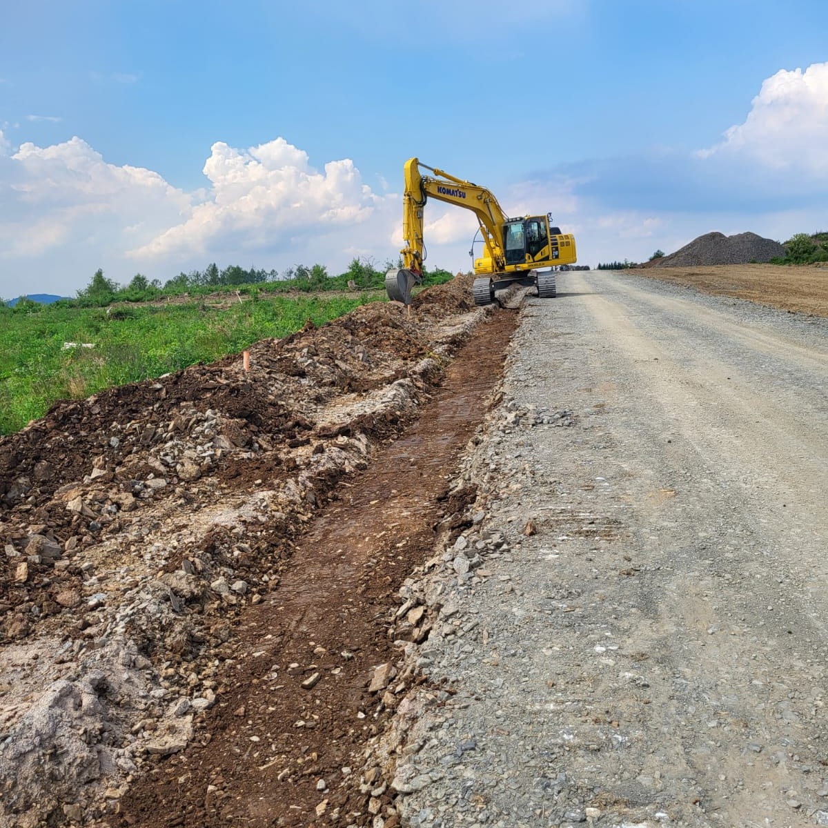 Żółta koparka Komatsu wyrównuje teren przy drodze gruntowej, widoczne ślady pracy i hałda ziemi w tle, błękitne niebo z białymi chmurami.