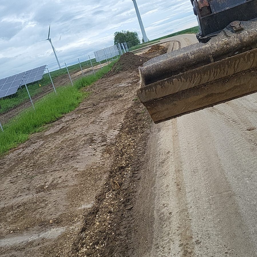 Łyżka koparki rozgarnia ziemię z kamieniami wzdłuż drogi, w tle panele słoneczne i turbiny wiatrowe.