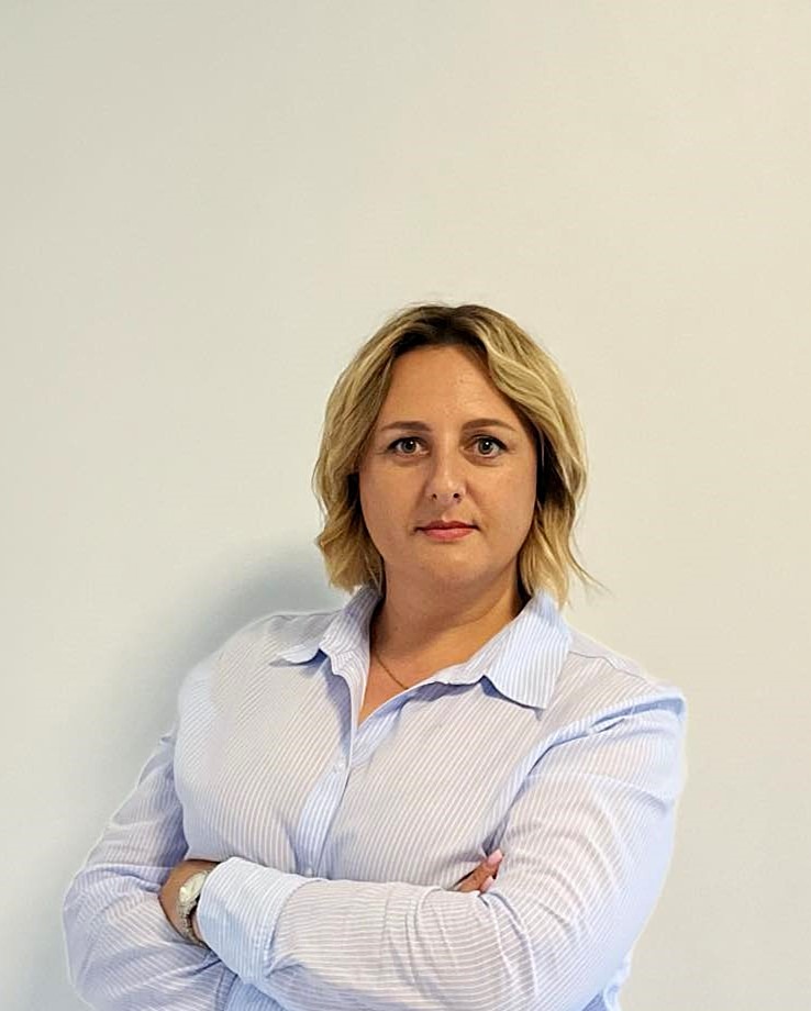 Portret kobiety w niebieskiej koszuli w paski, z rękami skrzyżowanymi na piersiach, patrzącej wprost w obiektyw na tle jasnej ściany.