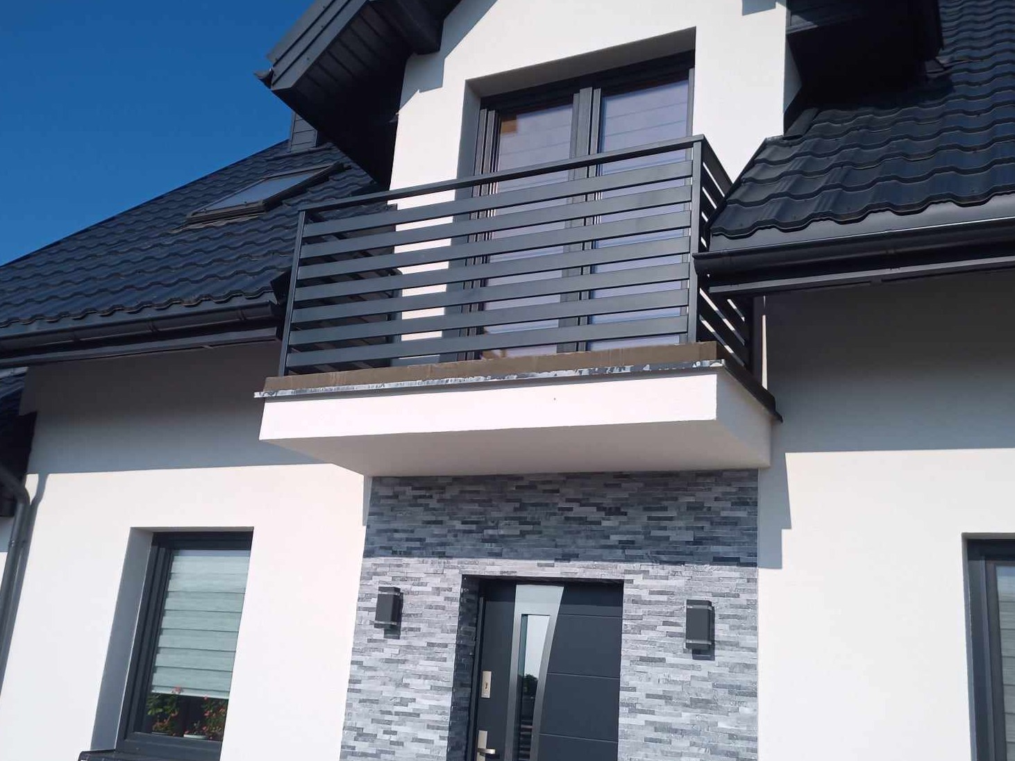 Nowoczesna, pozioma balustrada balkonowa w kolorze antracytowym, zamontowana na balkonie domu z elewacją w kolorze białym i szarym kamieniem dekoracyjnym.