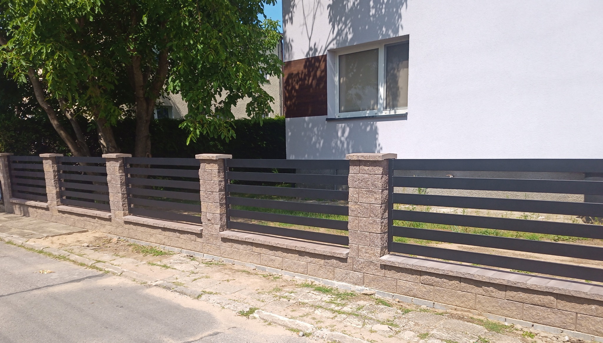 Nowoczesne ogrodzenie z poziomych, grafitowych przęseł metalowych osadzonych na słupkach i podmurówce z beżowej kostki łupanej, widok z zewnątrz posesji.