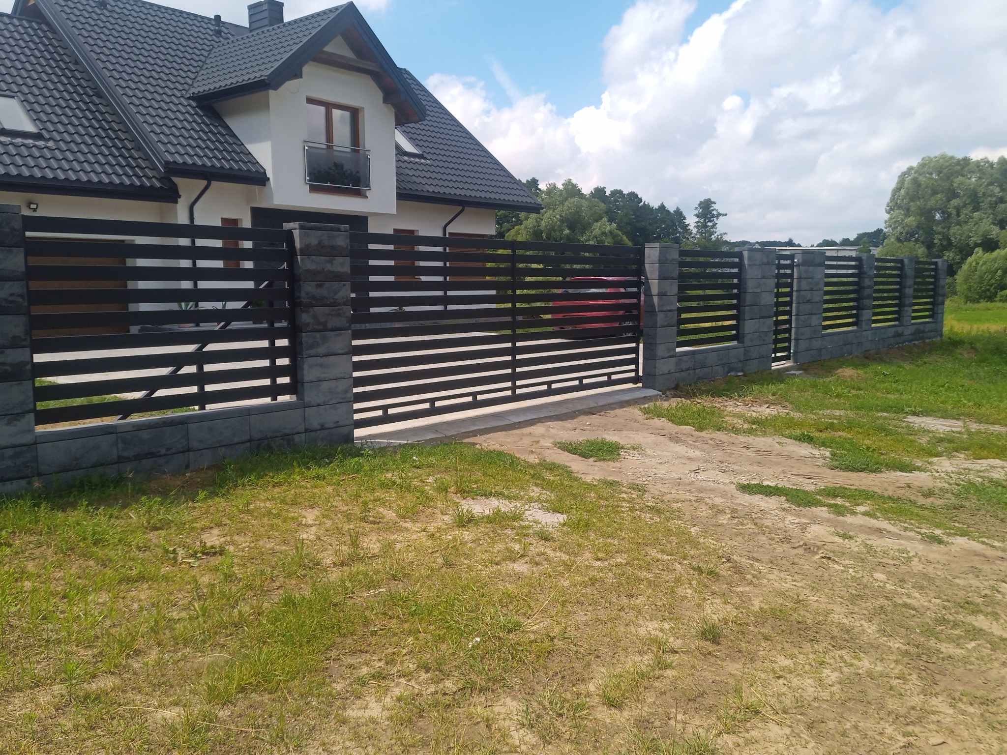 Nowoczesne ogrodzenie posesji z poziomych, ciemnych paneli zamontowanych między słupkami z szarego, łupanego kamienia; w tle widoczny dwukondygnacyjny dom z ciemną dachówką i fragment czerwonego...