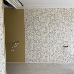 DNIPRO-VOLA SPÓŁKA Z OGRANICZONĄ ODPOWIEDZIALNOŚCIĄ - Pokój w trakcie tapetowania: jedna ściana z wzorzystą tapetą, druga pomalowana na jednolity kolor. Widoczne wystające kable elektryczne i listwy sufitowe.