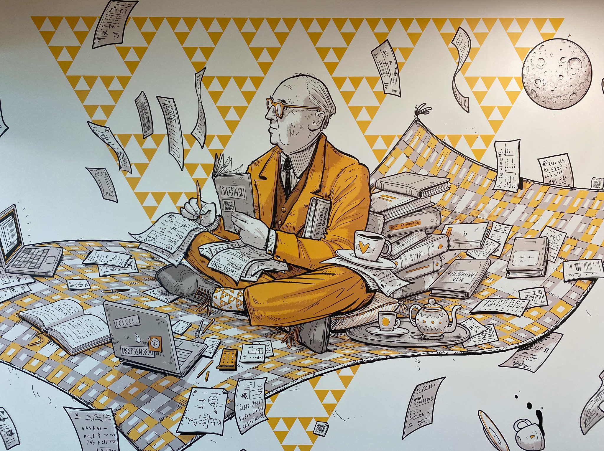 Ilustracja ścienna: Profesor w okularach czyta książkę 'Sierpiński' siedząc na kocu w otoczeniu książek, laptopów i notatek. W tle żółte trójkąty i księżyc.