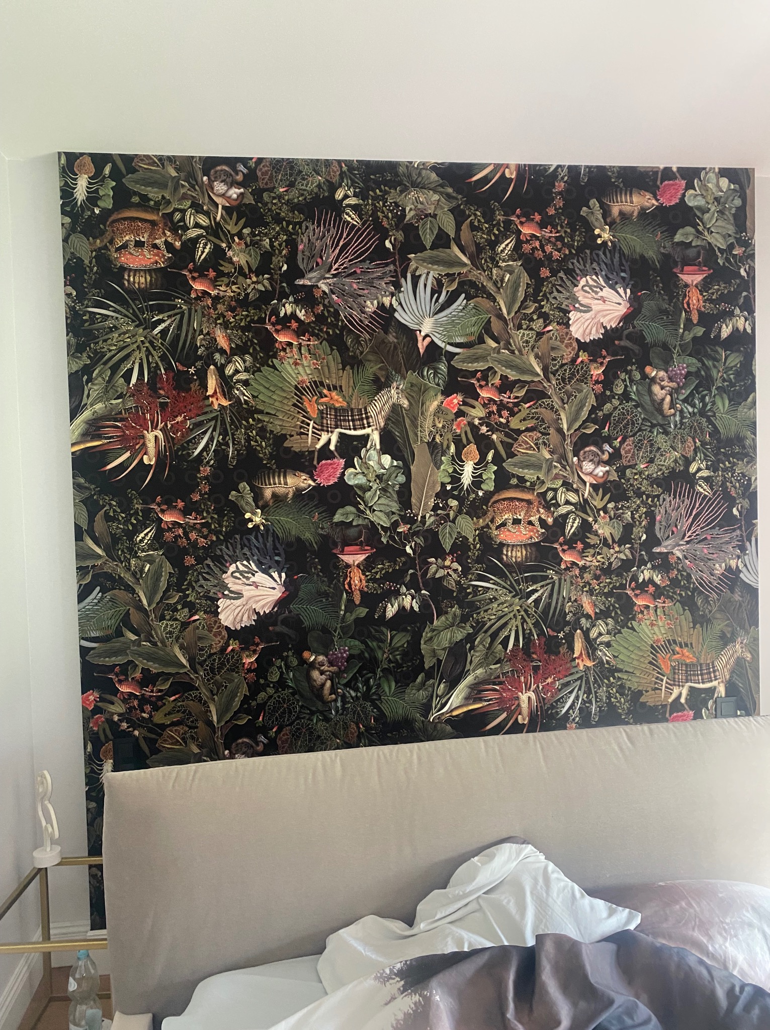 Ściana w sypialni pokryta tapetą w motywy dżungli z egzotycznymi zwierzętami i roślinami. Widoczny fragment łóżka z beżowym zagłówkiem. Dekoracja wnętrza.