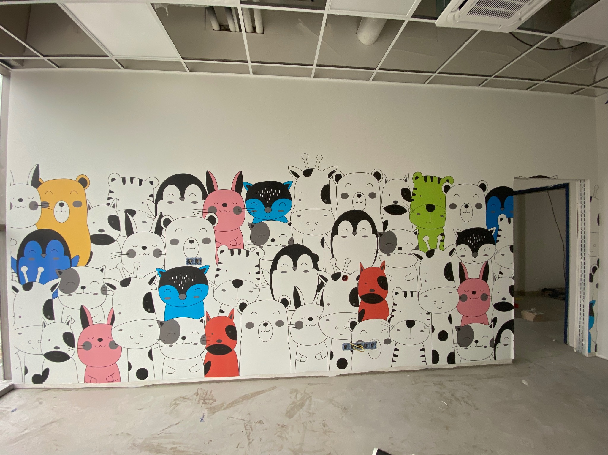 Ściana w biurze ozdobiona kolorowym, dziecięcym motywem zwierząt. Widoczne niedoskonałości w wykończeniu pod sufitem. Otwarta przestrzeń, surowy beton na podłodze.