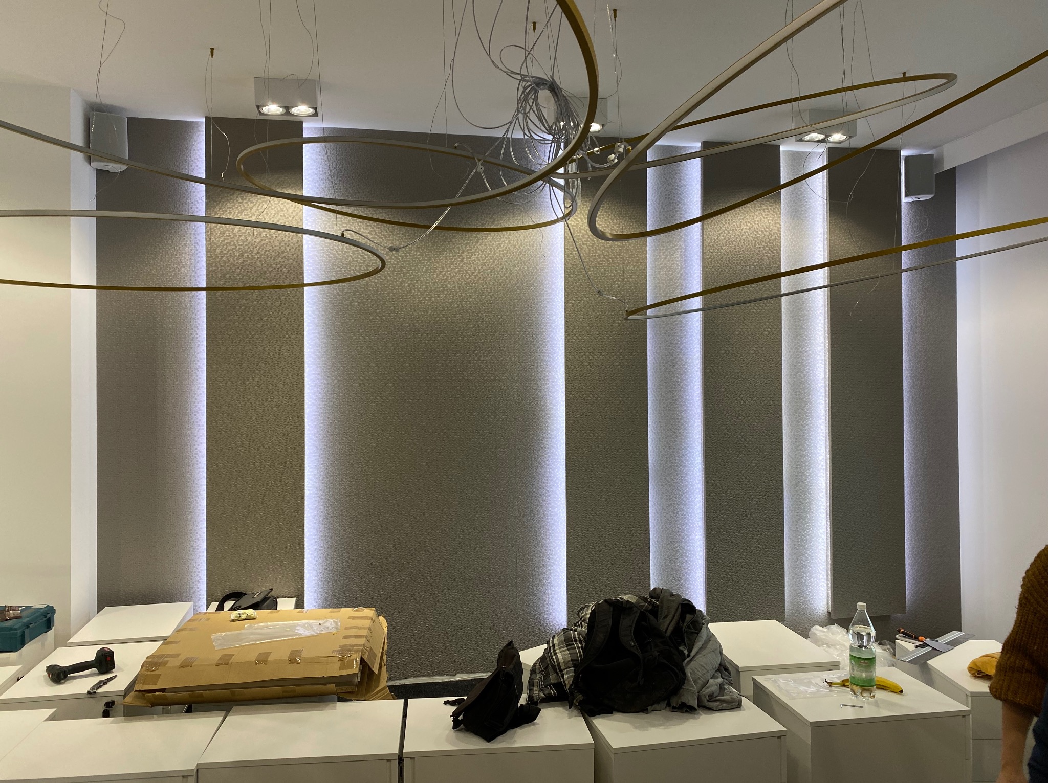 Wnętrze biura w trakcie wykańczania: tapeta w pionowe pasy, nowoczesne oświetlenie, narzędzia i materiały na białych szafkach. Widoczne kable i elementy instalacji.