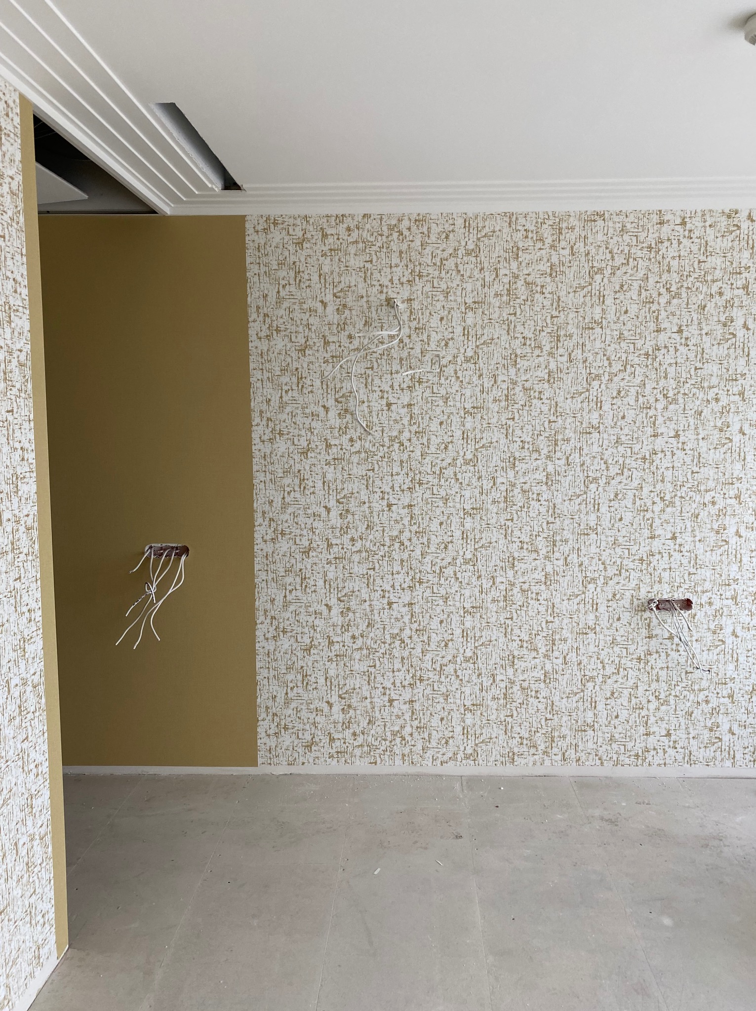 Pokój w trakcie tapetowania: jedna ściana z wzorzystą tapetą, druga pomalowana na jednolity kolor. Widoczne wystające kable elektryczne i listwy sufitowe.