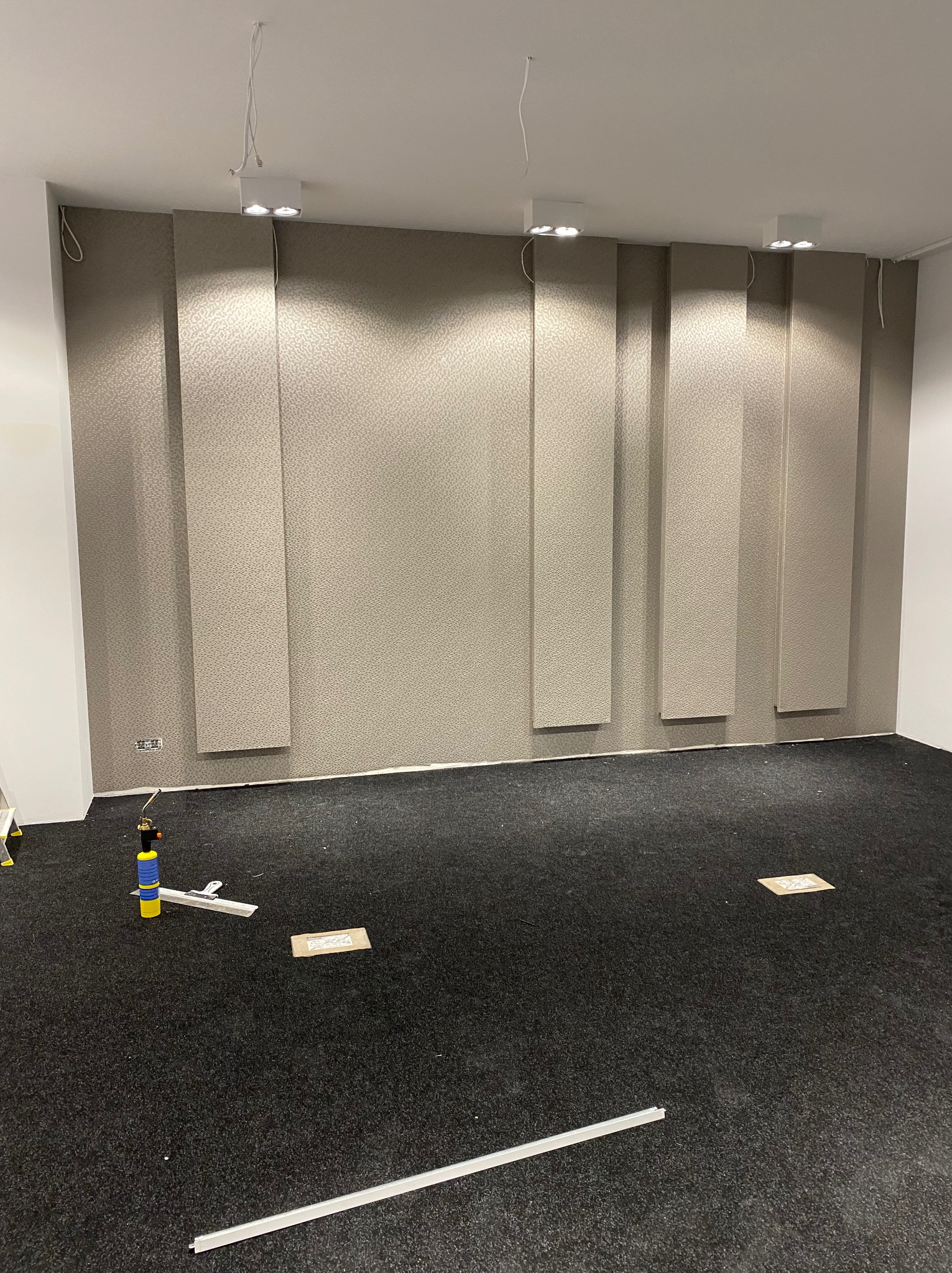 Wnętrze pokoju z częściowo wytapetowaną ścianą w odcieniach beżu. Widoczne narzędzia pracy, lampy sufitowe i czarna wykładzina podłogowa.