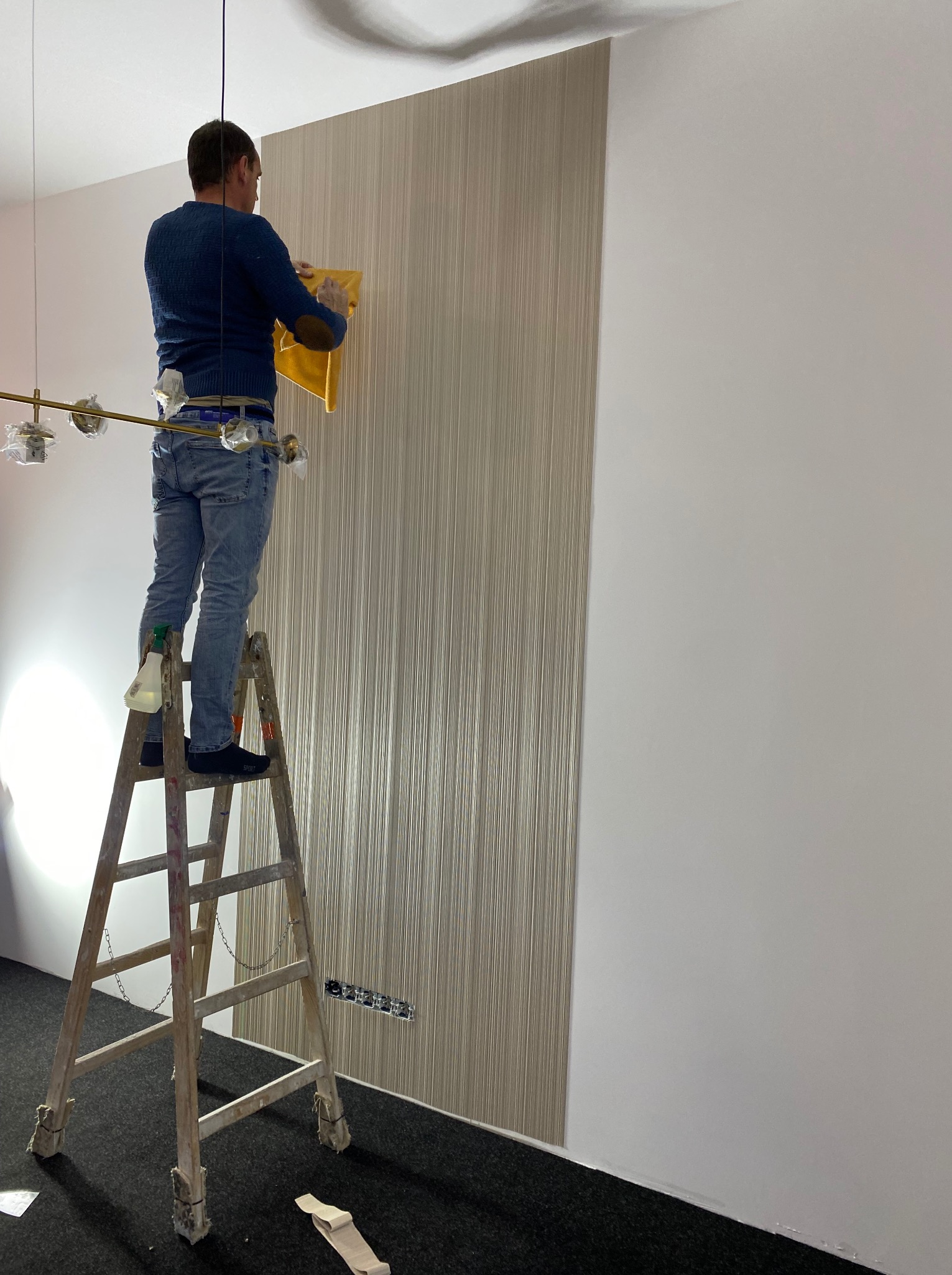 Pracownik na drabinie aplikuje tapetę w pionowe prążki na ścianie. Widoczny fragment ściany bez tapety, lampa sufitowa i narzędzia pracy.