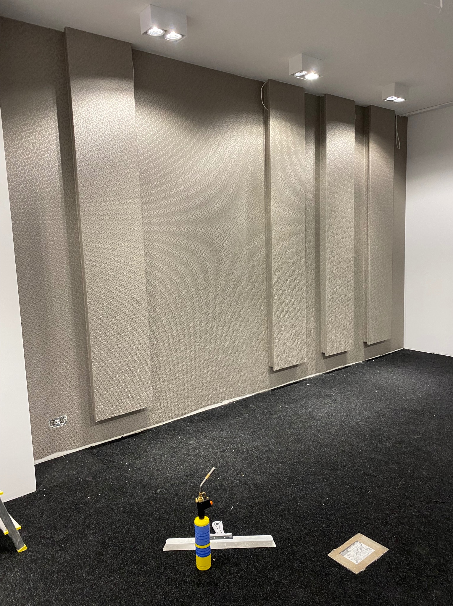 Wykończona ściana z dekoracyjnymi, pionowymi panelami tapetowanymi w odcieniach beżu. Na podłodze leży palnik i narzędzia, co sugeruje etap końcowy prac.