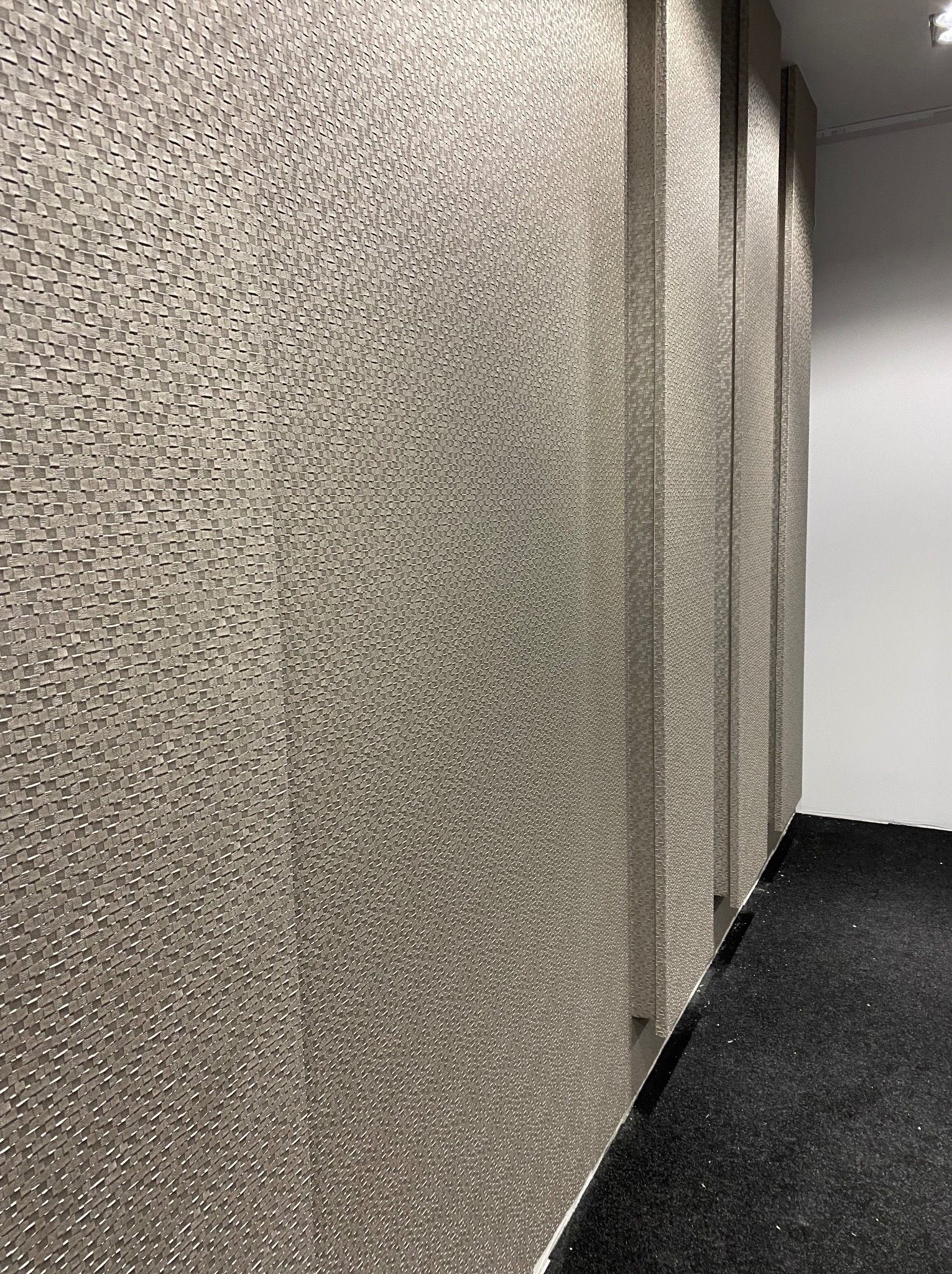 Teksturowana ściana z pionowymi panelami w odcieniach szarości, tworząca nowoczesny efekt dekoracyjny. Widoczny fragment podłogi z ciemną wykładziną.