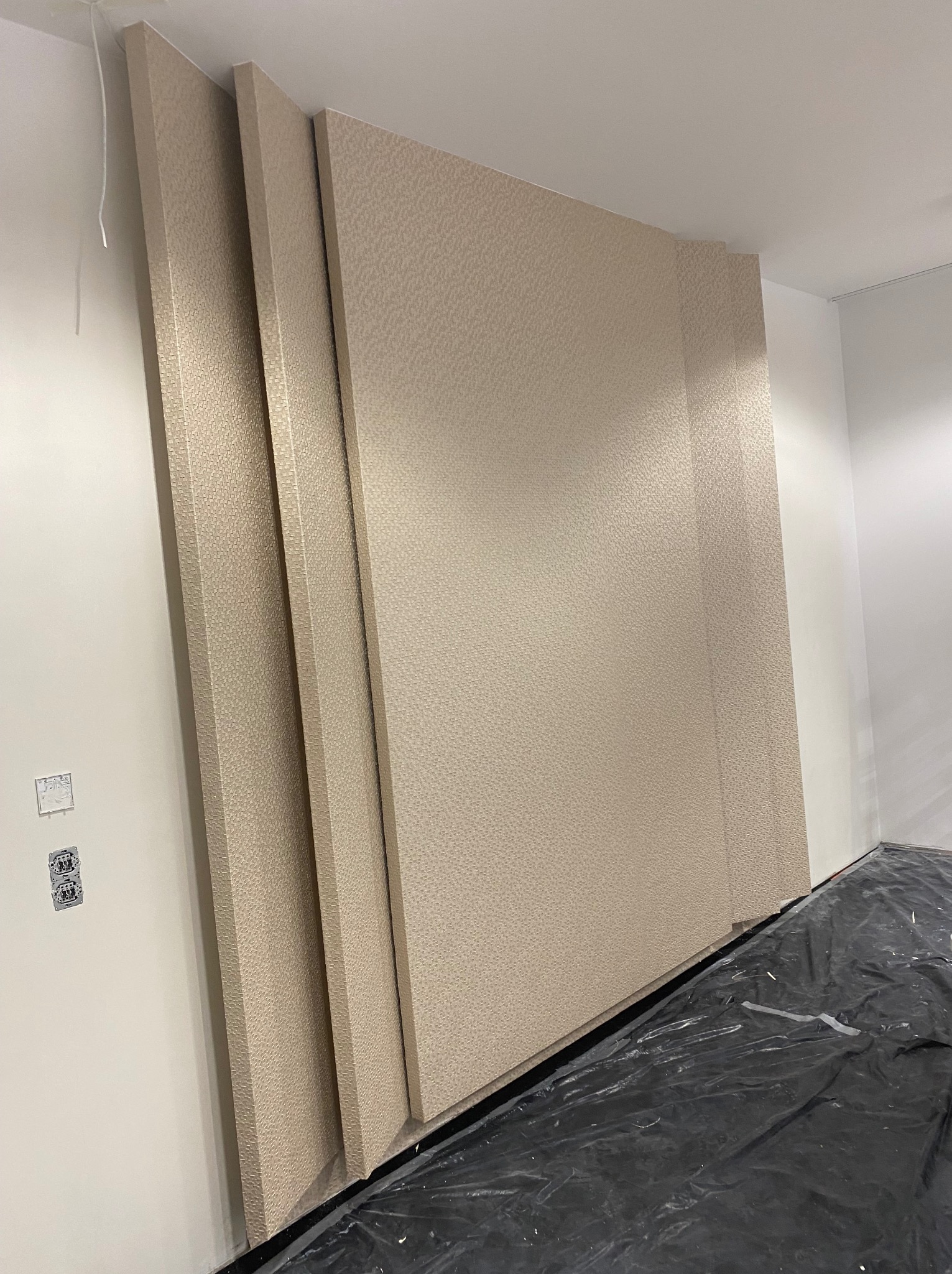 Beżowa tapeta z delikatnym wzorem na ścianie, widoczne fragmenty kilku pasów tapety, zabezpieczona folią podłoga, gniazdka elektryczne na ścianie.