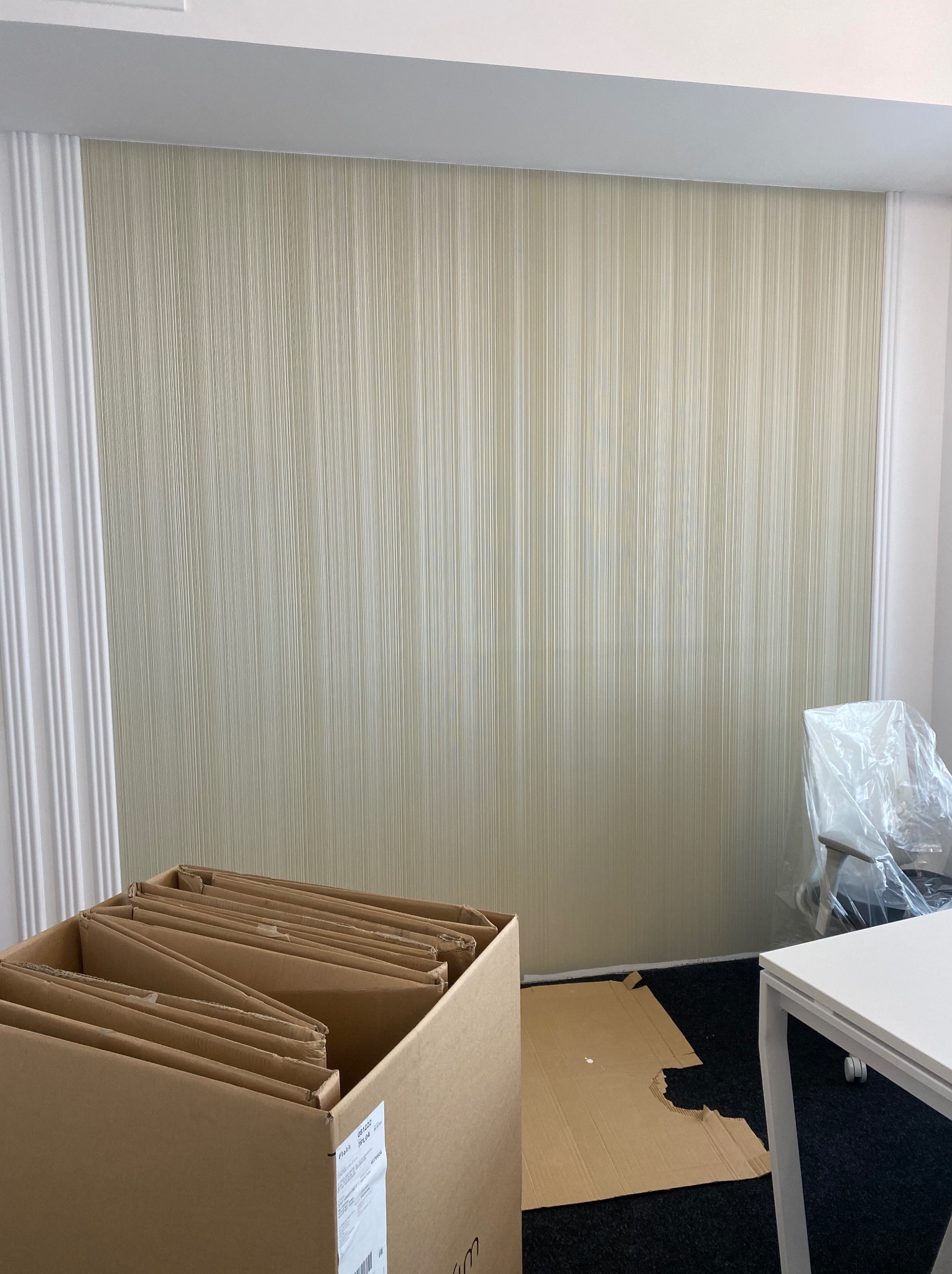 Ściana po tapetowaniu w biurze w Warszawie, z delikatną, pionową fakturą tapety. Widoczne kartony i zabezpieczone meble, co sugeruje świeżo zakończone prace.