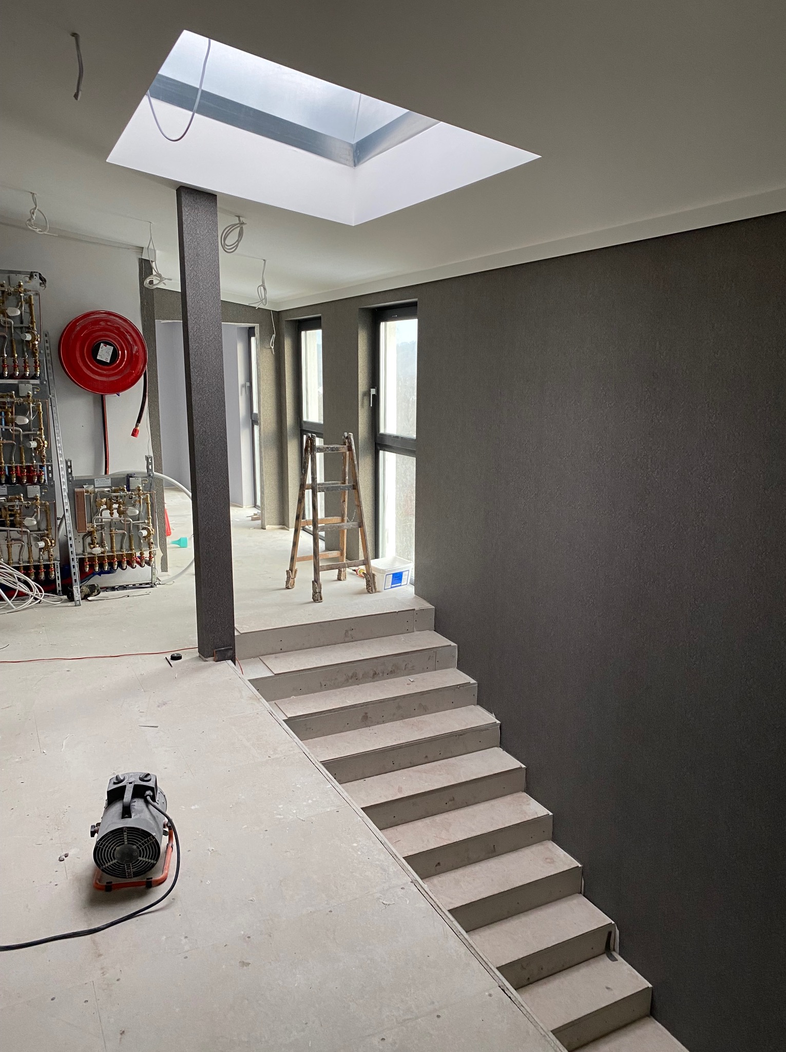 Klatka schodowa w trakcie prac wykończeniowych: ściana pokryta ciemną tapetą, surowe schody betonowe, drabina i sprzęt budowlany. Widoczne instalacje i okno dachowe.