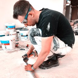 M&M - Pracownik w okularach ochronnych przycina metalowy profil szlifierką kątową akumulatorową na podłodze, w tle widoczne wiadra z farbą i inne narzędzia malarskie.