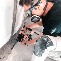 M&M - Pracownik w masce i okularach ochronnych, używa młota udarowego Dewalt do skuwania posadzki przy ścianie w trakcie remontu. Widoczne ślady po kuciu i zabrudzone ubranie robocze.