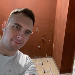 M&M - Selfie glazurnika w trakcie układania płytek podłogowych w łazience z pomarańczowymi ścianami. Widoczne krzyżyki dystansowe między płytkami imitującymi drewno.