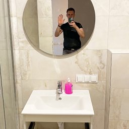 M&M - Wykończona łazienka z umywalką i lustrem, na ścianach płytki imitujące marmur. W lustrze odbija się osoba robiąca zdjęcie.