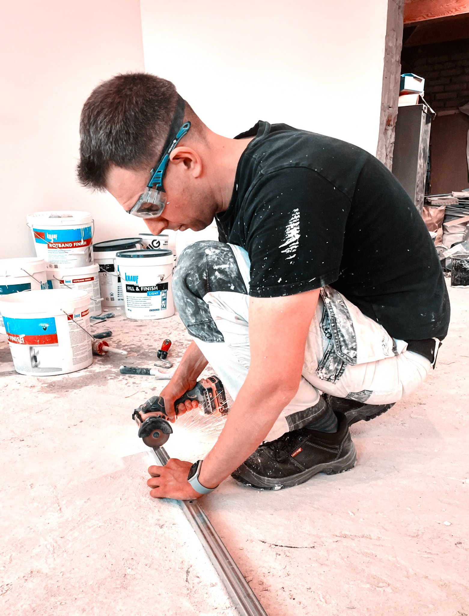 Pracownik w okularach ochronnych przycina metalowy profil szlifierką kątową akumulatorową na podłodze, w tle widoczne wiadra z farbą i inne narzędzia malarskie.