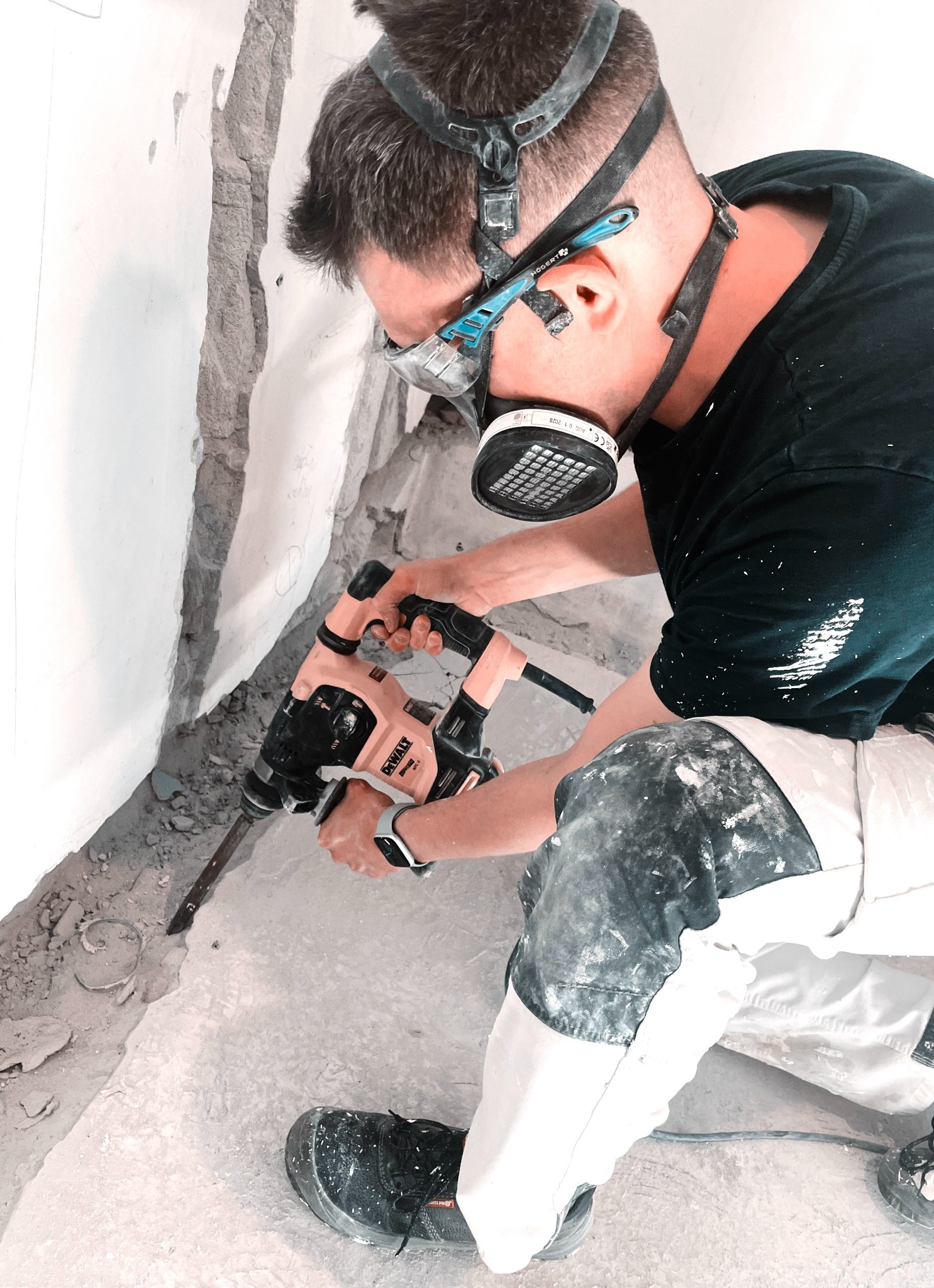 Pracownik w masce i okularach ochronnych, używa młota udarowego Dewalt do skuwania posadzki przy ścianie w trakcie remontu. Widoczne ślady po kuciu i zabrudzone ubranie robocze.