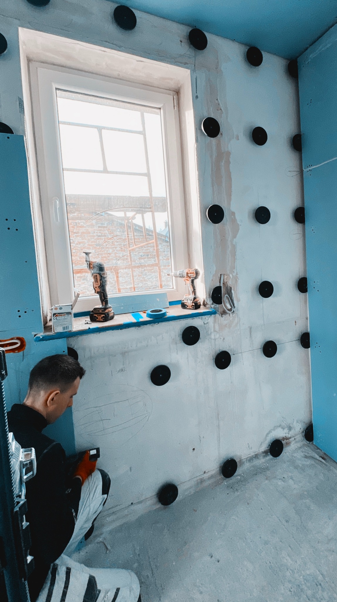 Pracownik w trakcie przygotowywania ściany w łazience do położenia płytek, widoczne czarne kołki mocujące, narzędzia na parapecie okna, rysunek na ścianie.