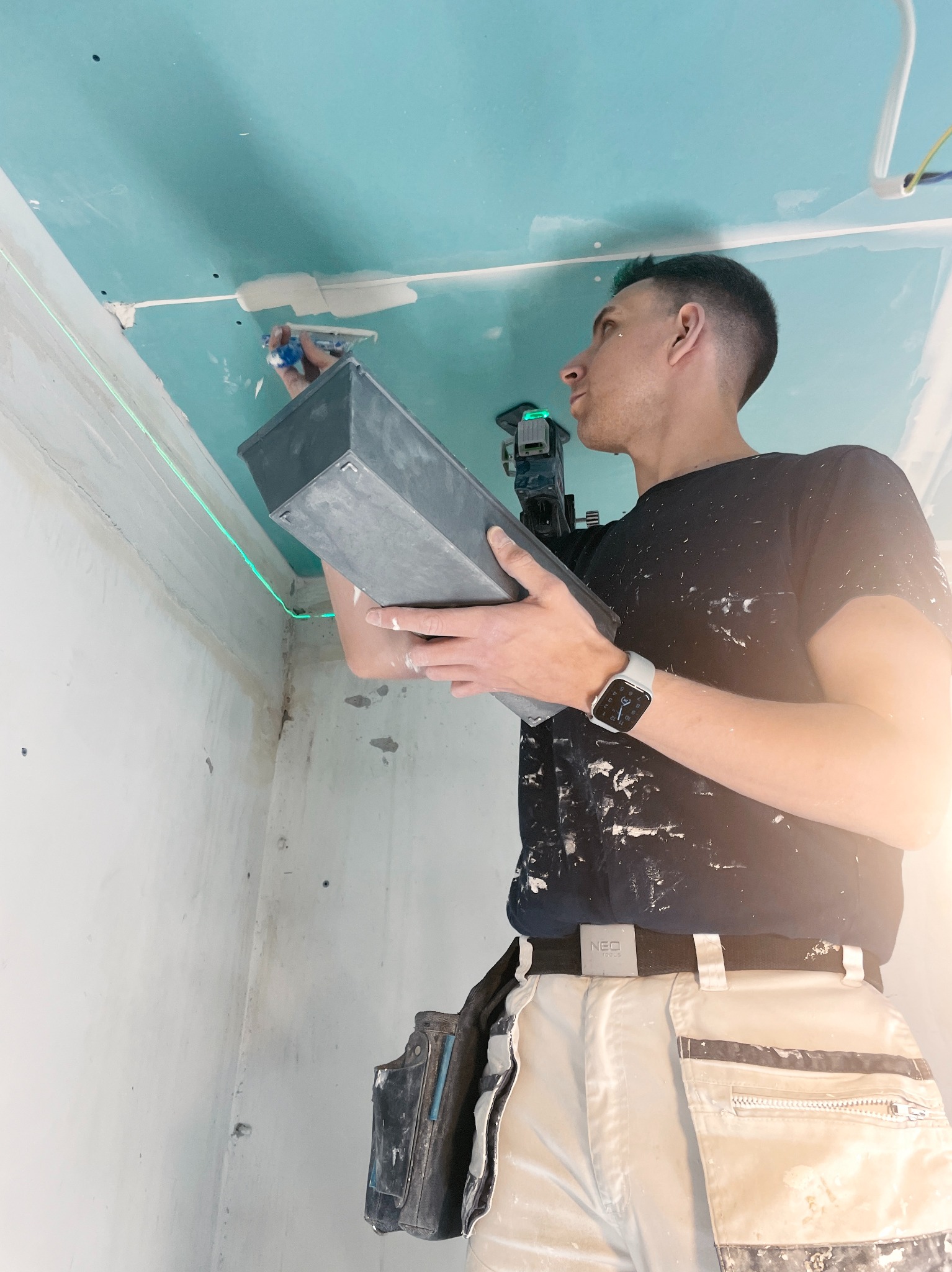 Glazurnik wyrównuje sufit z płyty gipsowej, używając szpachelki i lasera krzyżowego, widoczny na wysokości wzroku, z ubrudzonymi farbą spodniami roboczymi marki NEO.