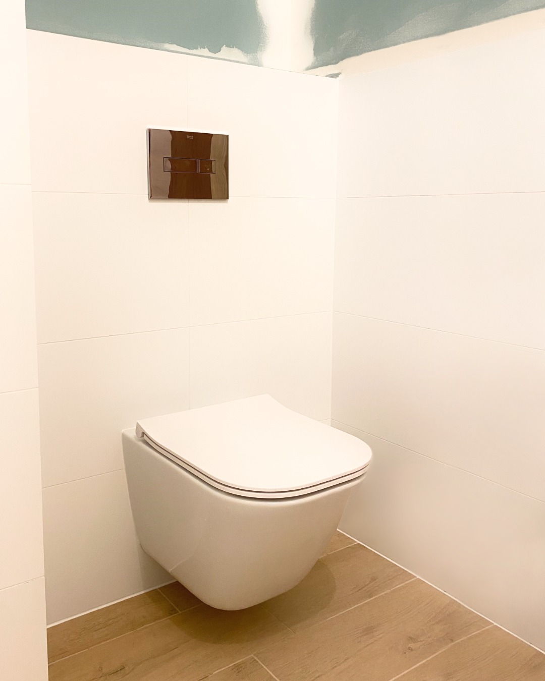 Podwieszana toaleta z brązowym przyciskiem spłukującym na białych płytkach ściennych, podłoga wyłożona panelami imitującymi drewno, surowy sufit w tle.