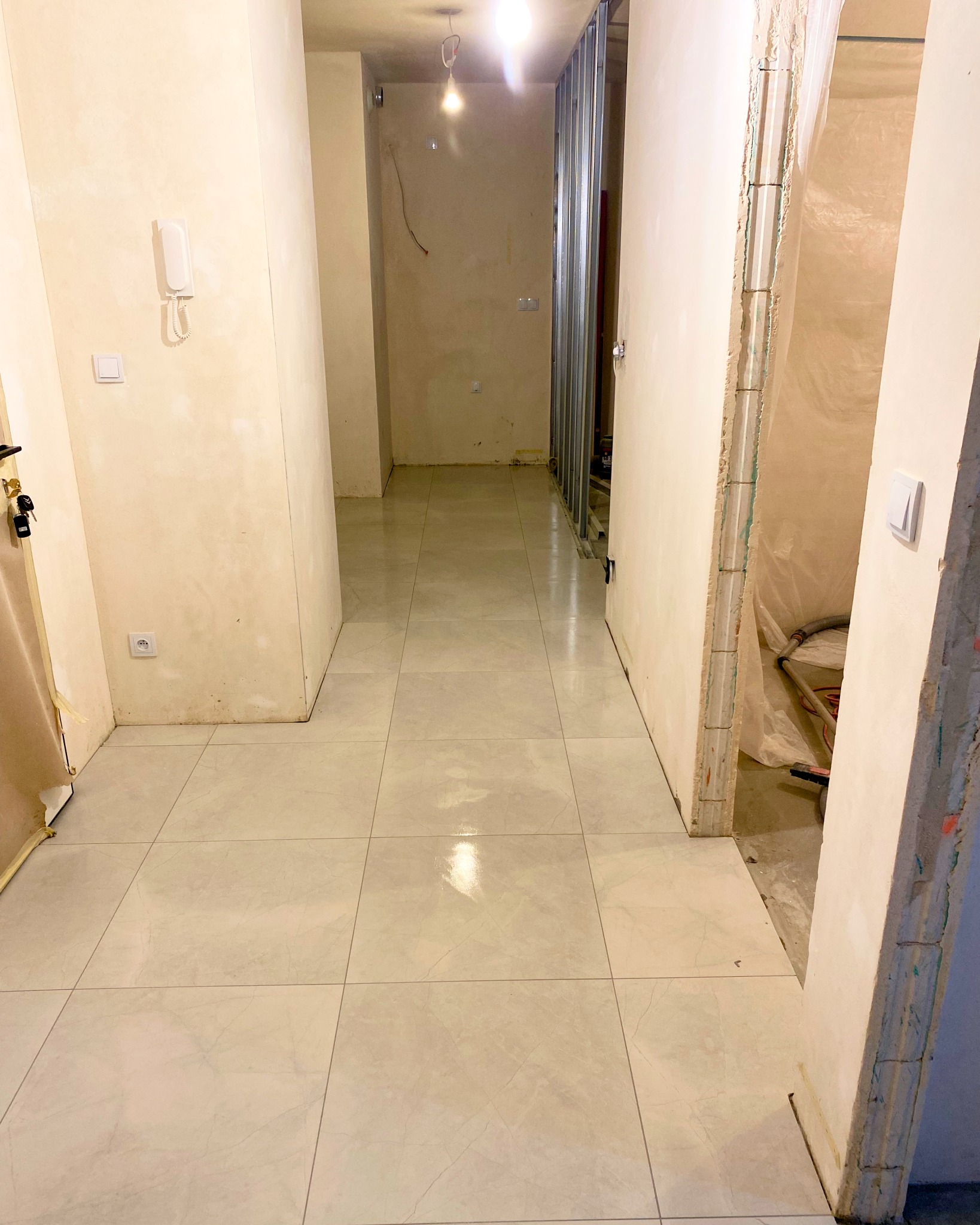 Korytarz w trakcie remontu z położonymi jasnymi, kwadratowymi płytkami podłogowymi, widoczne surowe ściany i metalowa konstrukcja ścianki działowej.