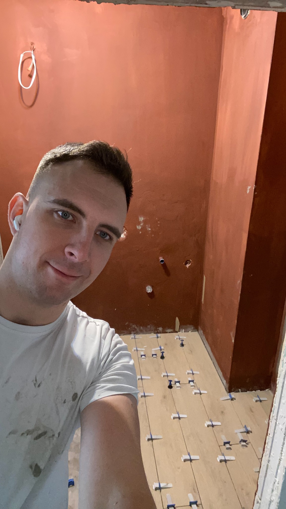 Selfie glazurnika w trakcie układania płytek podłogowych w łazience z pomarańczowymi ścianami. Widoczne krzyżyki dystansowe między płytkami imitującymi drewno.