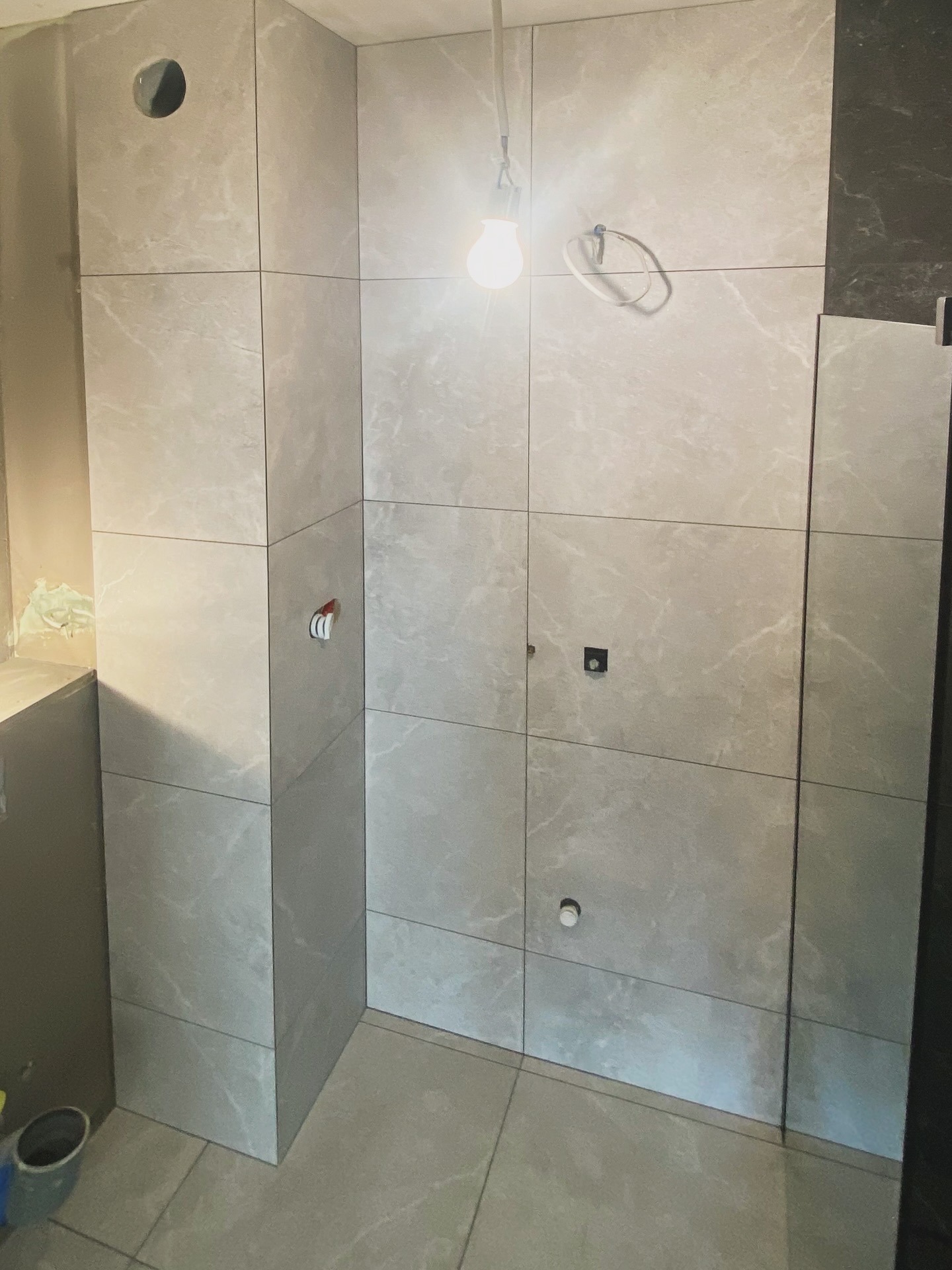 Wykończona ściana łazienki z jasnymi, marmurkowymi płytkami. Widoczne otwory na instalacje, surowy stan ściany obok i wisząca żarówka.