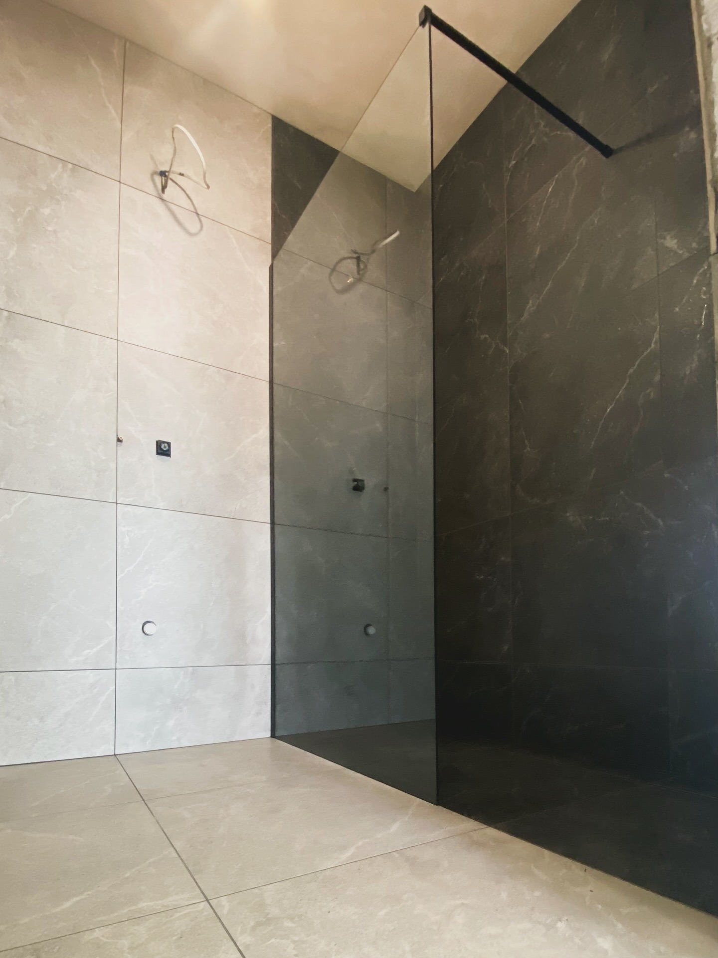 Częściowo wykończona łazienka z szarymi płytkami imitującymi marmur i szklaną kabiną prysznicową z czarnymi ramami. Widoczne przygotowane instalacje.